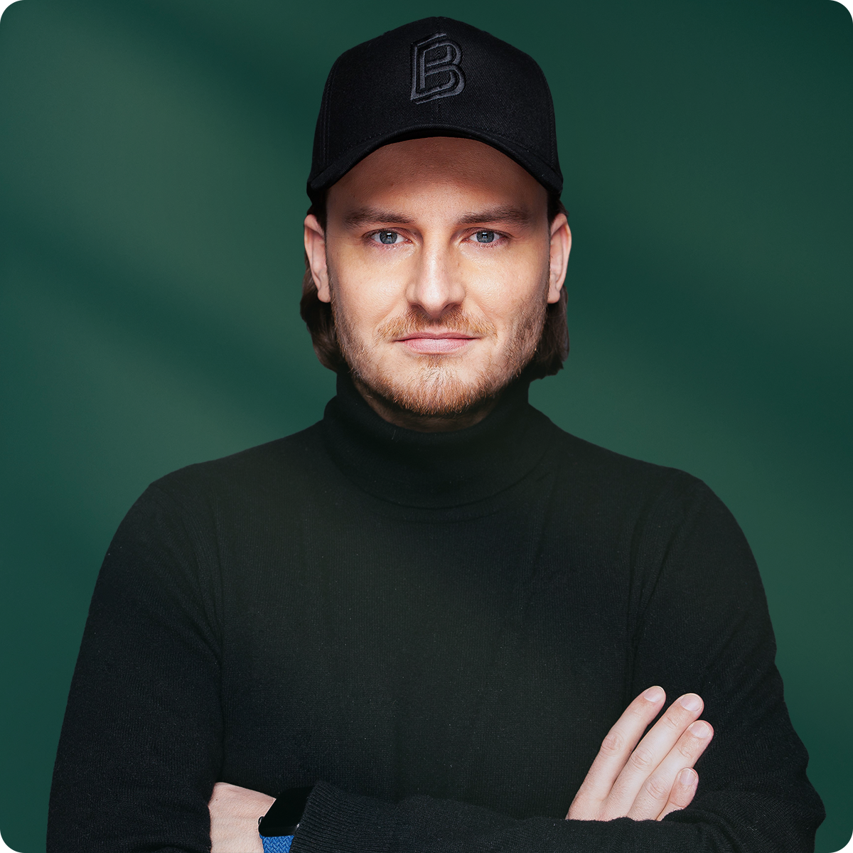 Man in a black cap and turtleneck with arms crossed, standing against a green background.
