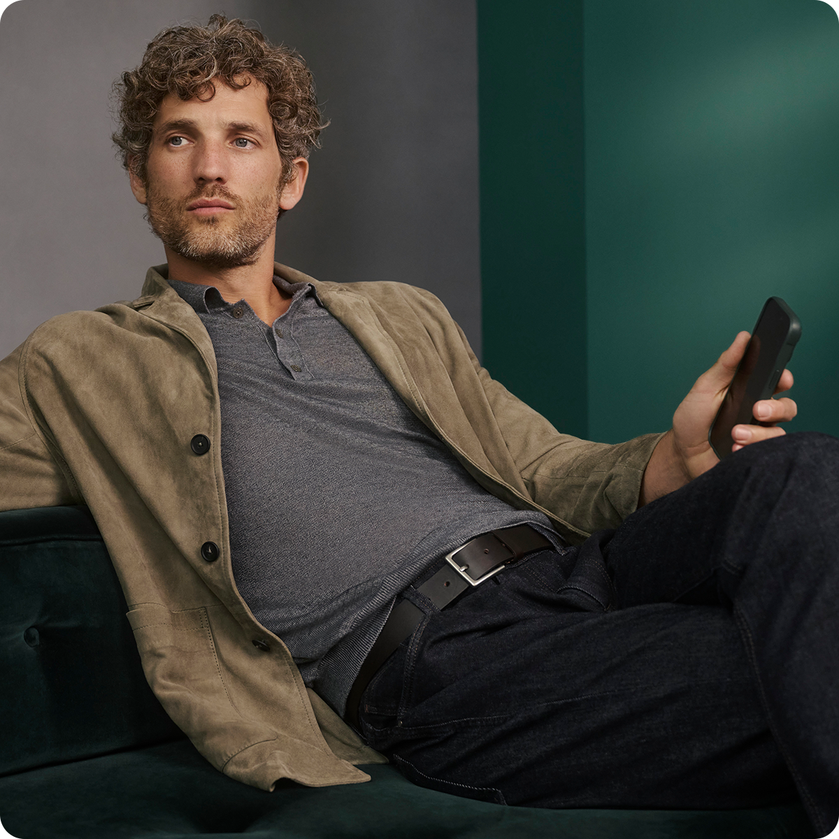 Man with curly hair in a beige jacket and gray shirt, holding a smartphone, reclines on a dark green sofa against a green and gray backdrop.
