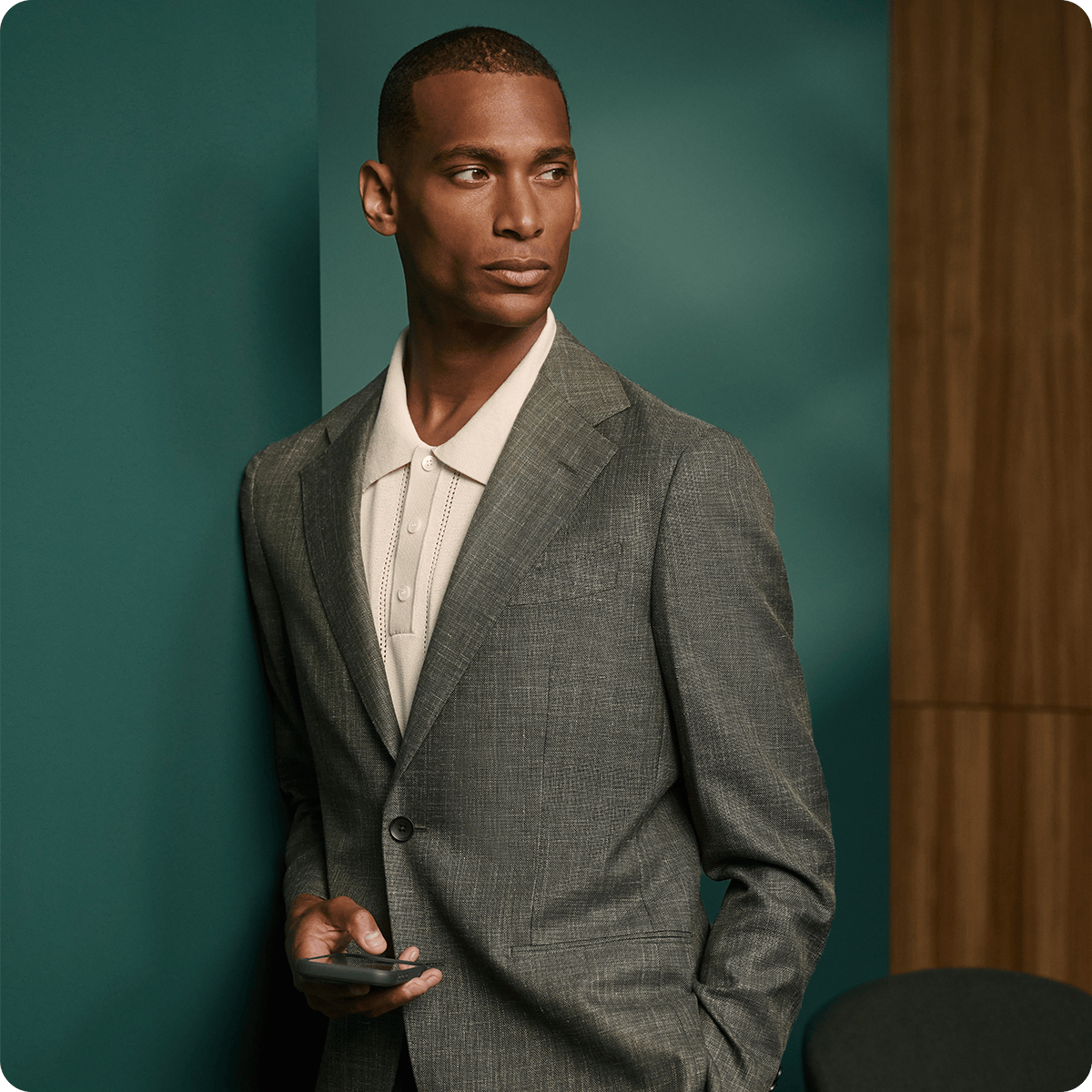 Man in a gray suit and white shirt holding a phone, standing against a green and wood-paneled background, looking to the side.
