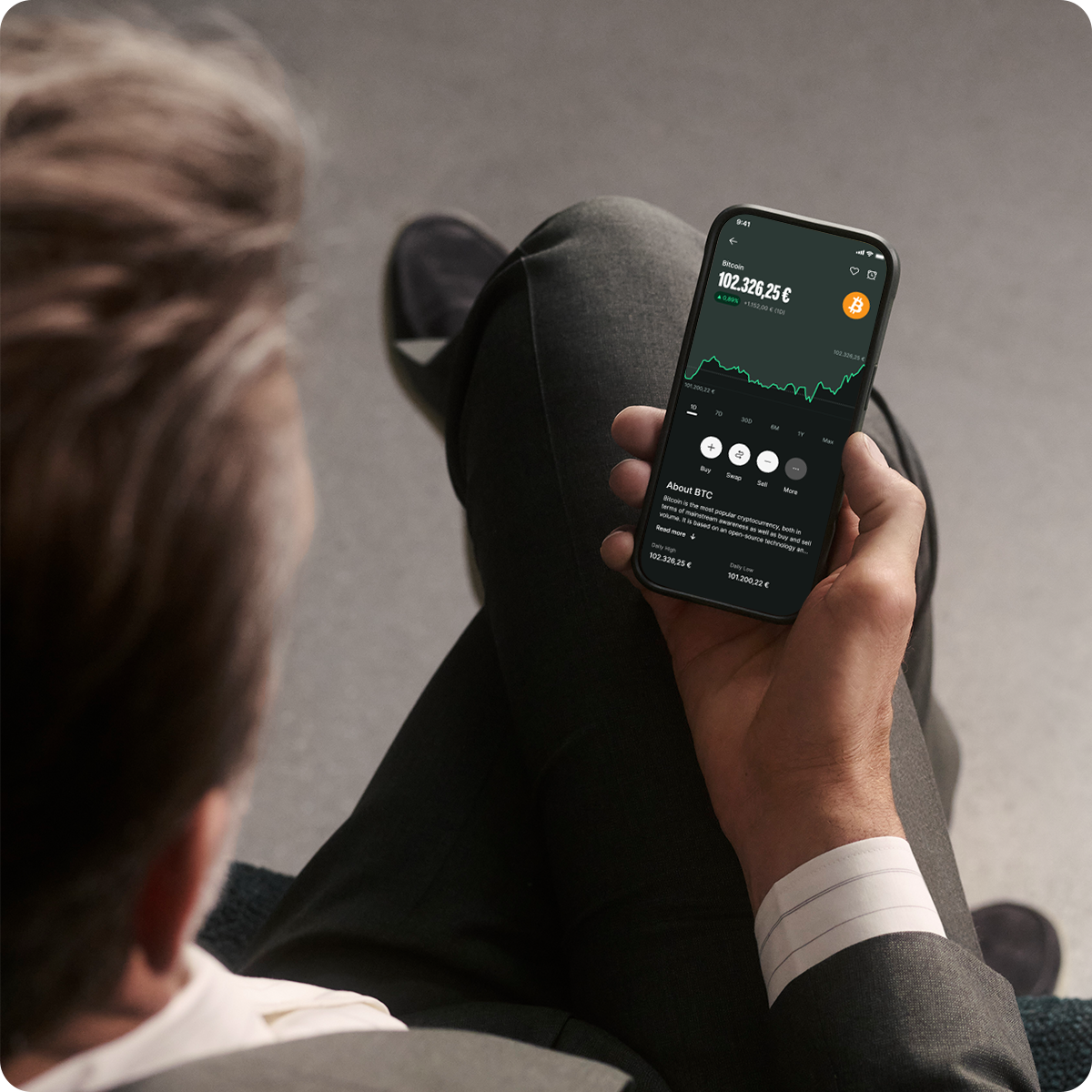 Person in a suit holding a smartphone displaying a cryptocurrency trading app with graphs and data.